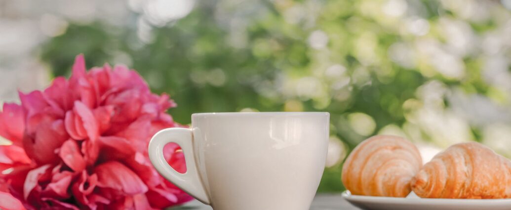Ceașcă de cafea alături de un mic dejun cu pâine, ouă și fructe, pe o masă luminată de soare