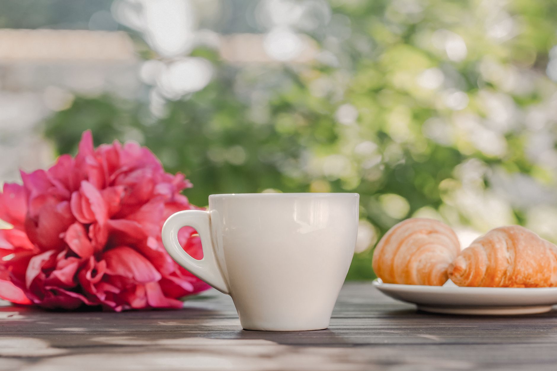Ceașcă de cafea alături de un mic dejun cu pâine, ouă și fructe, pe o masă luminată de soare